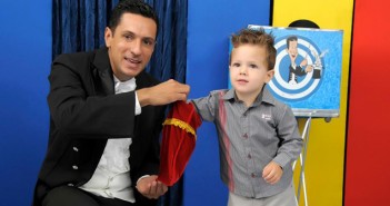 Oficina de mágicas, dia 21 de fevereiro Mágico Ulisses Loddy segura um saquinho de veludo vermelho enquanto uma criança de aproximadamente 3 anos coloca a mão dentro para puxar alguma coisa.