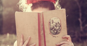 Um livro por semana? Menina loira, com laço vermelho na cabeça, lê um livro com capa bege. O livro esconde o seu rosto.