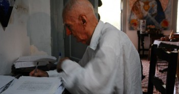 Um ano sem Suassuna O escritor Ariano Suassuno, de perfil, em sua escrivaninha, escrevendo em folhas de papel. Ao lado dele, uma pilha de folhas.